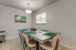 Un coin repas comprenant une table, des chaises, un luminaire au plafond, une fenĂȘtre et un tableau encadrĂ© accrochĂ© au mur.