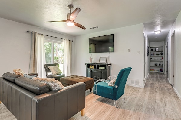 A living room with a sofa, ottoman, teal armchair, fireplace, TV, and a hallway leading to a closet. - 7 Mi to Six Flags! Family Retreat in DFW Suburbs (Irving)