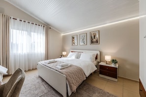 Ein Schlafzimmer mit einem Bett, Nachttischen, einem Sessel und einem Fenster mit Vorhängen.