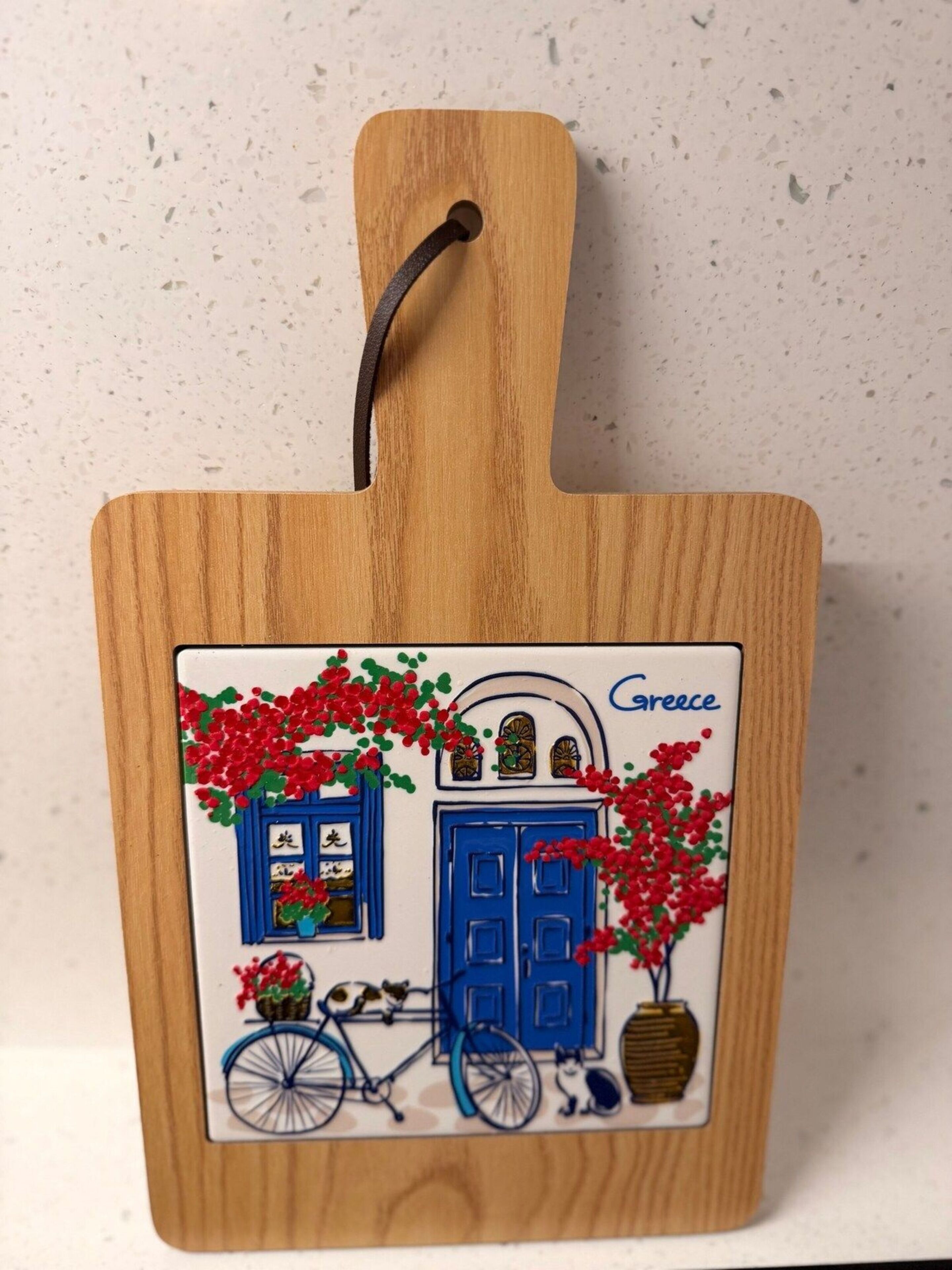 Una tabla de madera con un asa colgante, que presenta una escena pintada de una casa azul con flores rojas, una bicicleta y un gato.