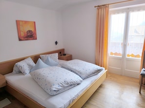 A double bed with floral bedding, a bedside table with a lamp, a wall clock, and a door with curtains.