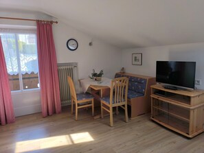 A room with a wooden table, two chairs, a TV on a wooden stand, a radiator, a clock, and a window with curtains.