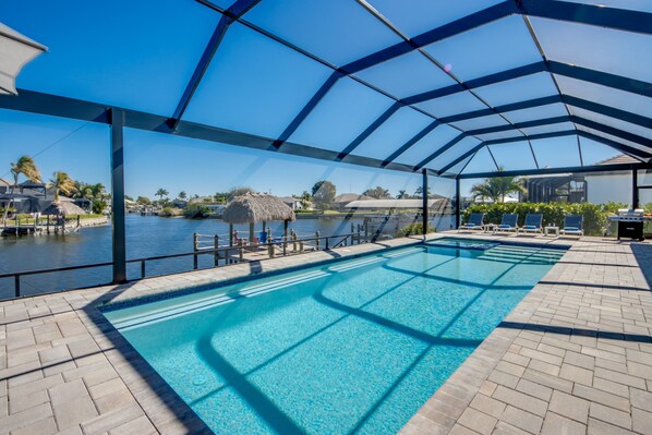 A covered swimming pool with clear blue water, a view of a body of water, and a thatched-roof structure in the background. - Villa Happy Place - on water, pool, spa, views, boating (Cape Coral)
