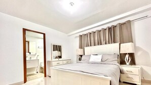 A bedroom with a bed, bedside tables, a nightstand, a mirror, and a door leading to a bathroom. - Penthouse 2BR w/ Private Rooftop Terrace + Views (Santo Domingo)