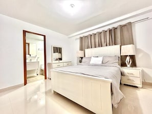 Une chambre à coucher comprenant un lit, des tables de chevet, une commode, un miroir et une porte donnant accès à une salle de bain.