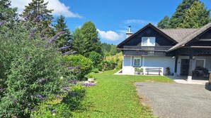Une maison avec une allée en gravier, une terrasse en bois et un jardin agrémenté de fleurs violettes.