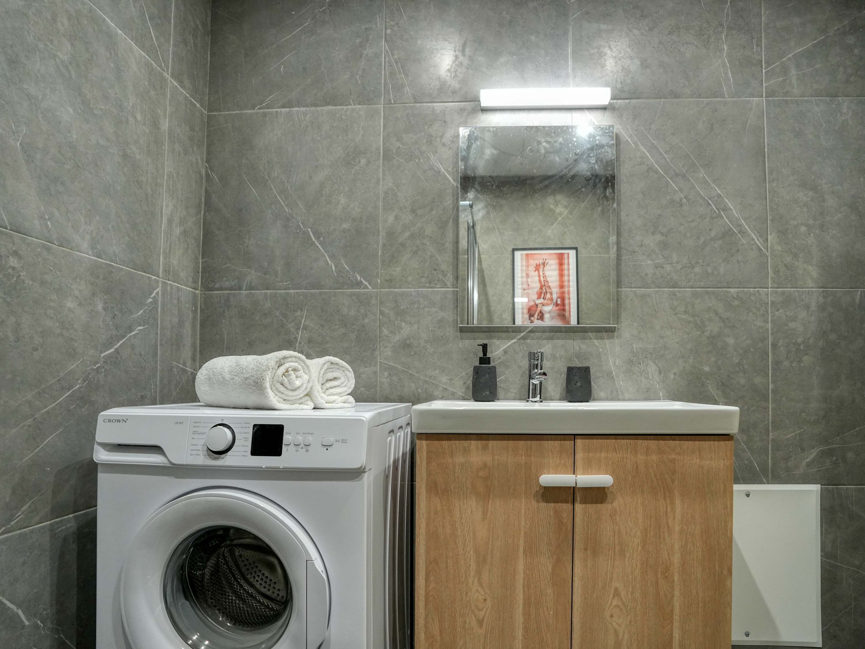 A modern bathroom with grey tiles, a washing machine, a wooden cabinet, and a framed picture on the wall.