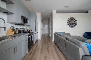 A modern kitchen with grey cabinets, a microwave, and a stove. There is a wooden countertop with knives and a plant. A grey sofa with blue cushions is visible in the background. - Dallashaus Pool Gym Love Field (Dallas)