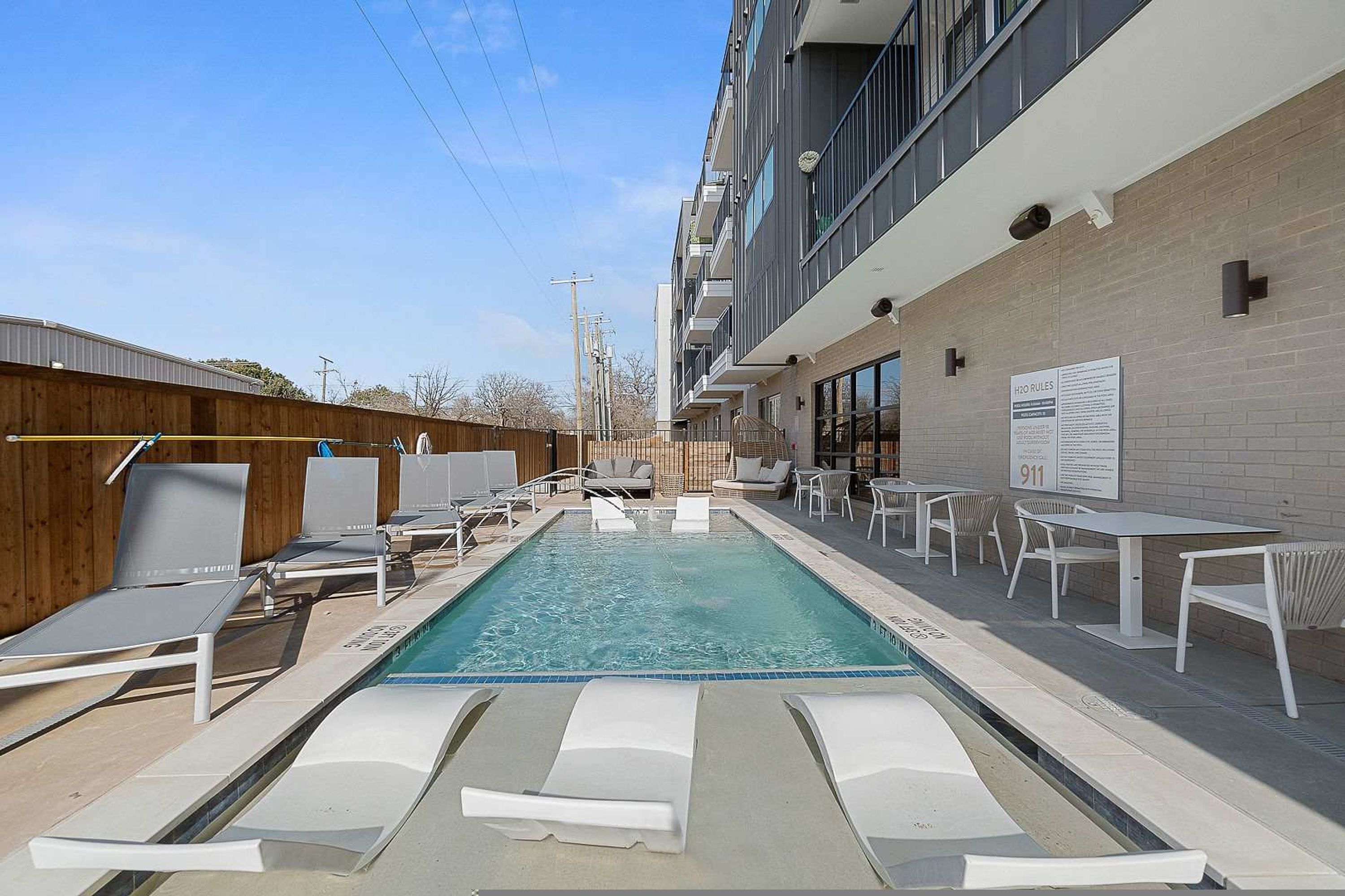 Un área de piscina con sillas de descanso y mesas, un cartel con normas y un edificio con balcones.