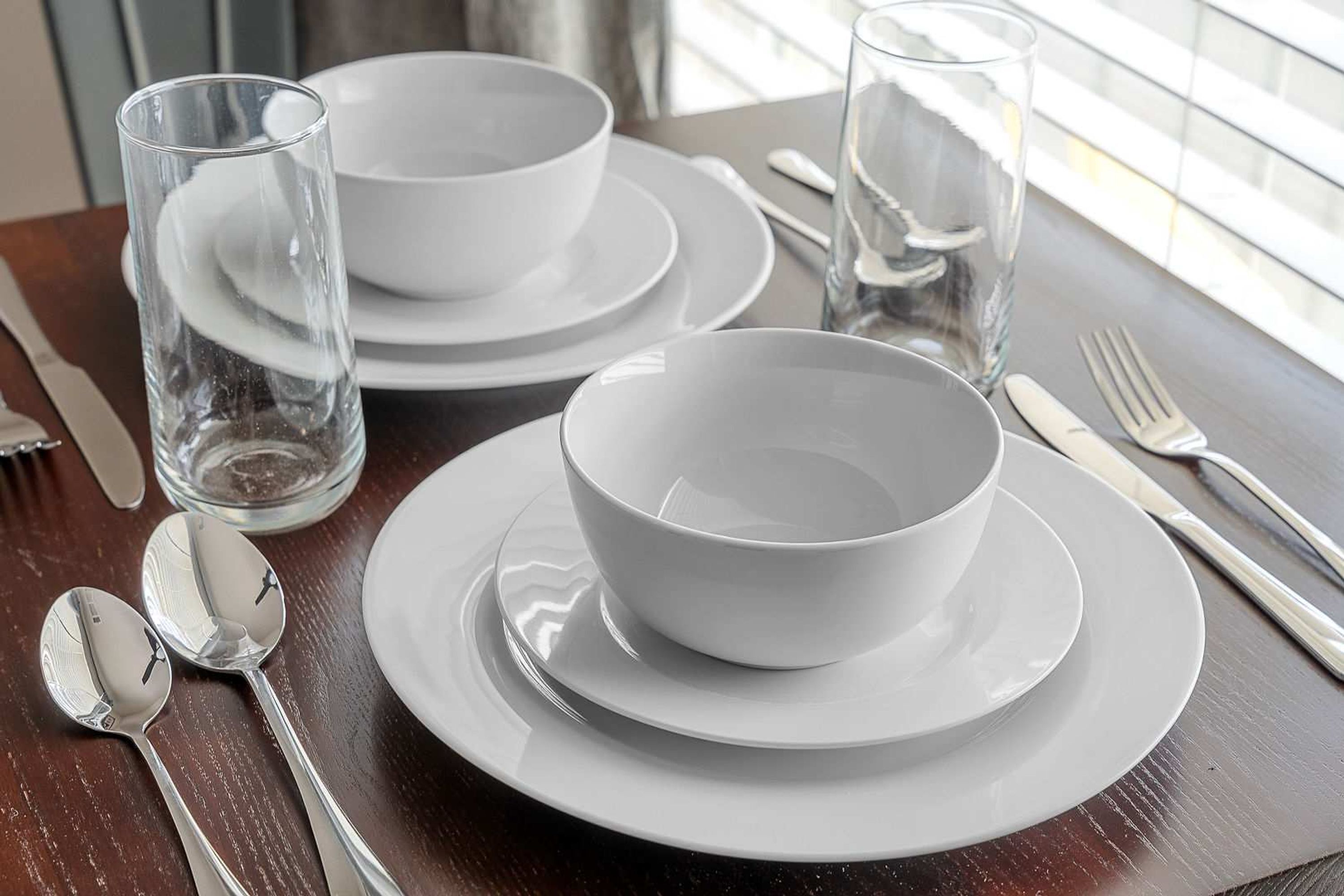 Una mesa dispuesta con platos, cuencos, cubiertos y vasos blancos sobre una superficie de madera.
