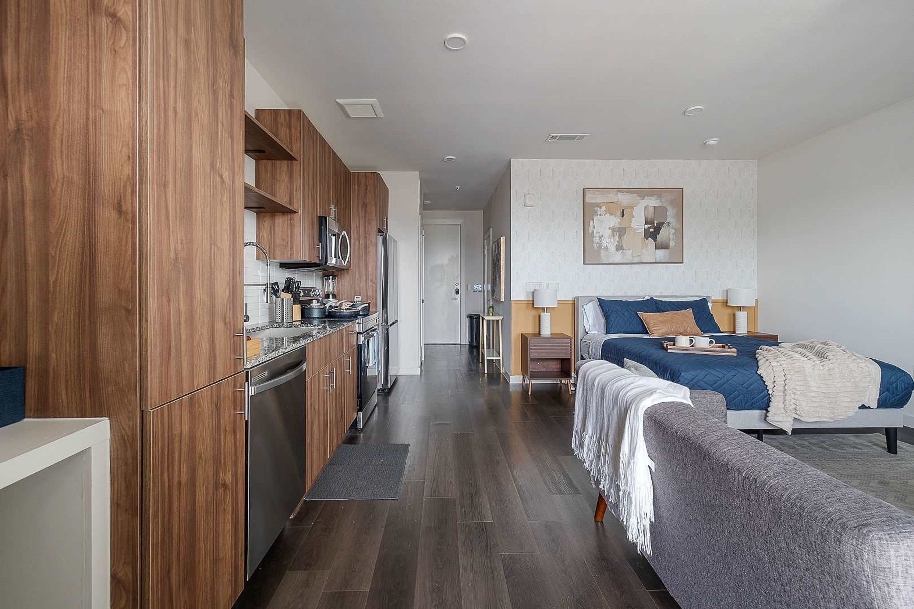 A modern hotel room with a kitchenette, a bed with a blue quilt, a bedside table, and a wall-mounted artwork.