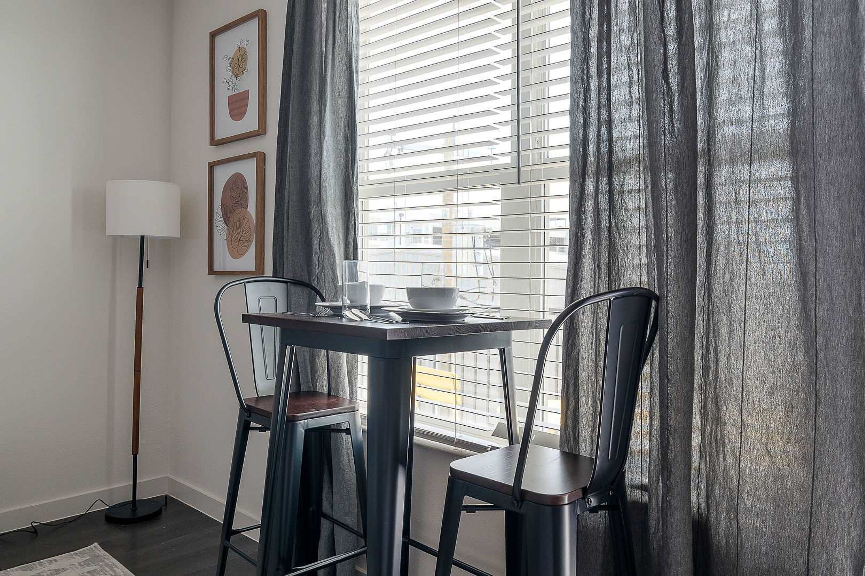 A dining area with a table and two chairs, a floor lamp, and two framed pictures on the wall.