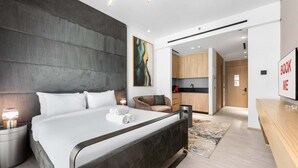Une chambre d’hôtel moderne avec un grand lit, un bureau, une chaise et un coin cuisine visible à travers une porte ouverte.