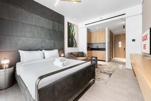 Ein modernes Hotelzimmer mit einem großen Bett, einem Schreibtisch, einem Sessel und einer durchgehenden Tür erkennbaren Küchenzeile.