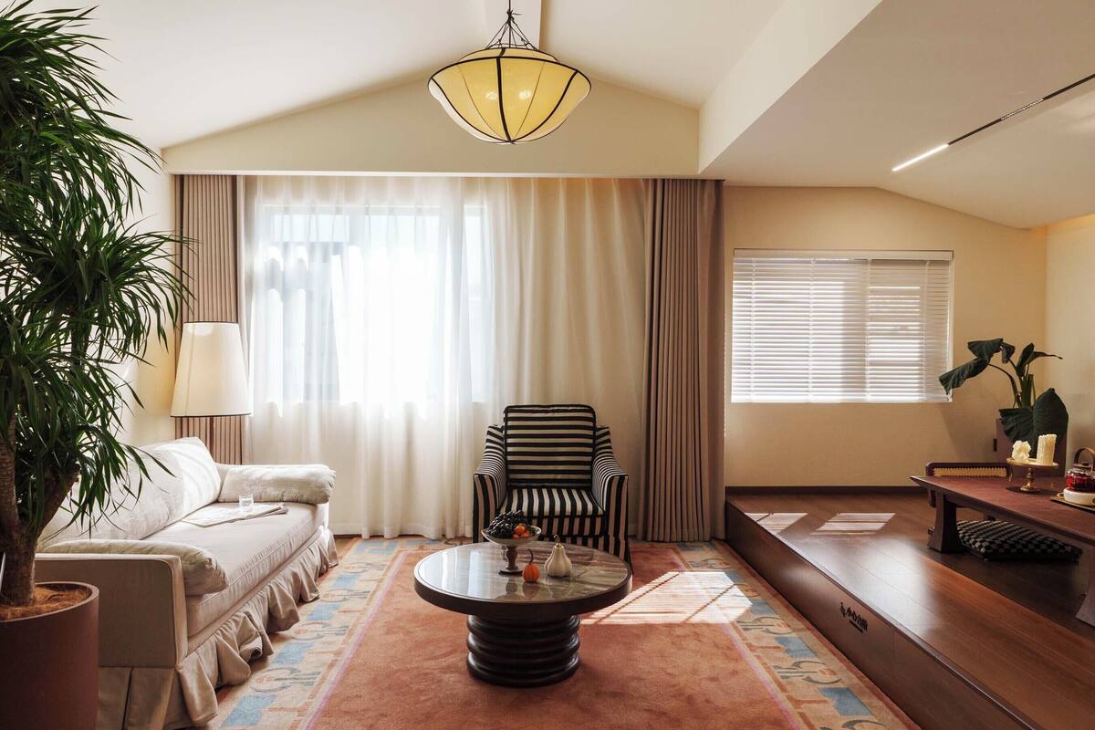 Quarto de hotel com sofá, poltrona listrada, mesa de centro redonda, luminária, planta em vaso e vista para o exterior através de amplas janelas.