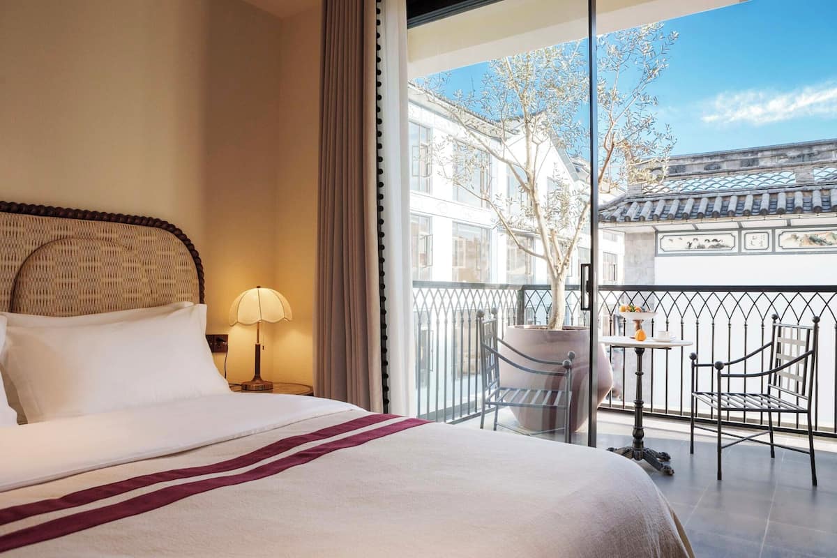 Quarto de hotel com cama, mesa de cabeceira, luminária, varanda com cadeiras e vista para arquitetura tradicional.