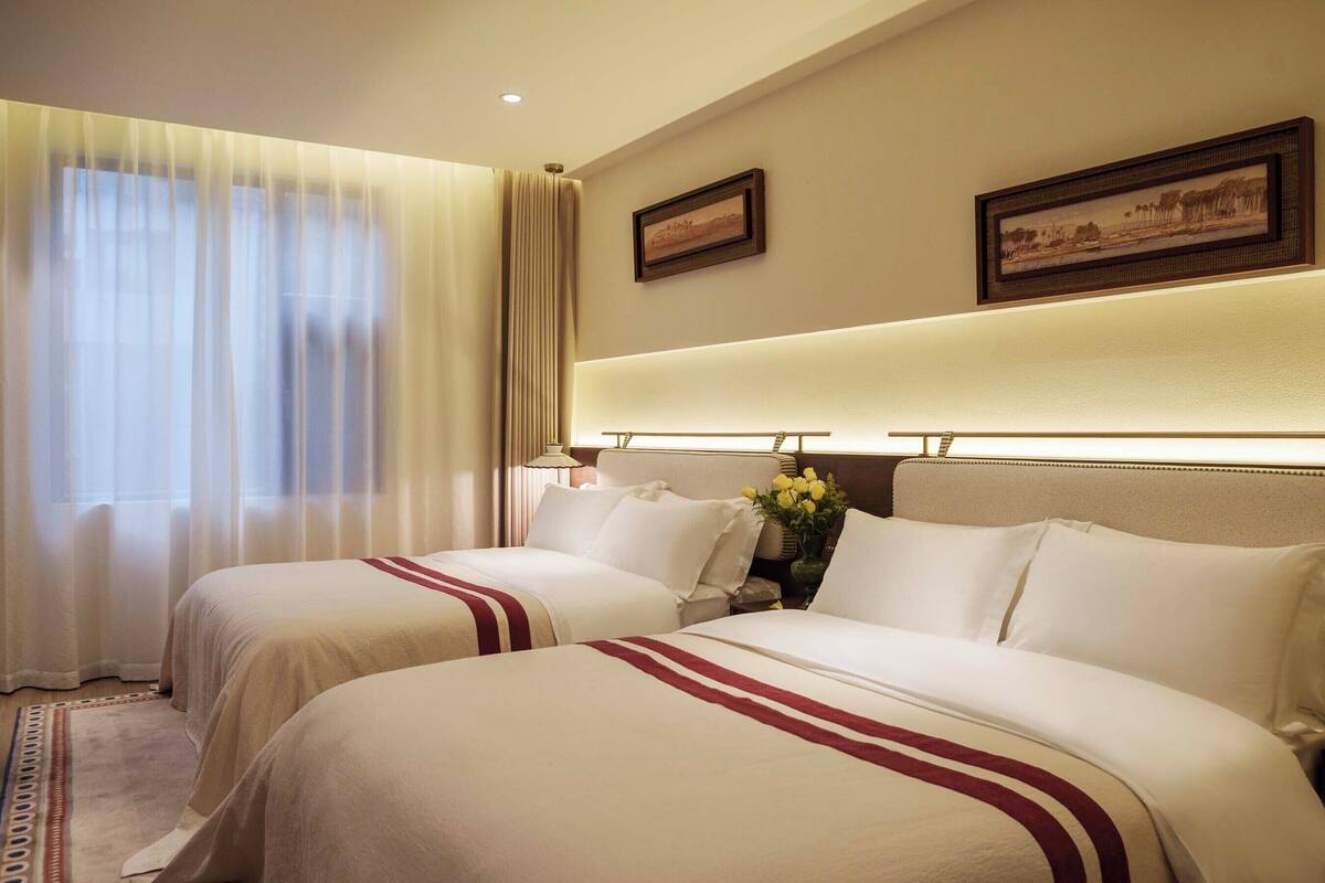 Quarto de hotel com duas camas, uma janela grande com cortinas, cabeceira com luz e dois quadros na parede.