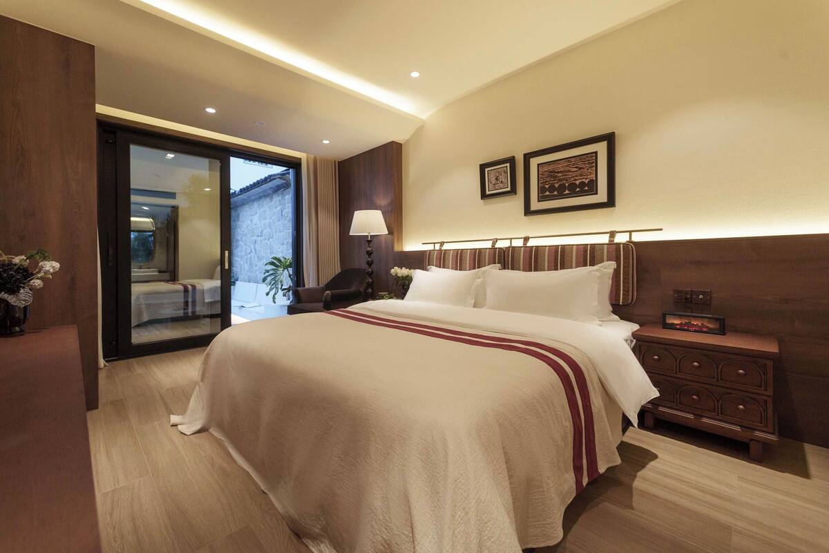 Quarto de hotel com uma cama grande, mesa de cabeceira, abajur e vista por uma porta de vidro.