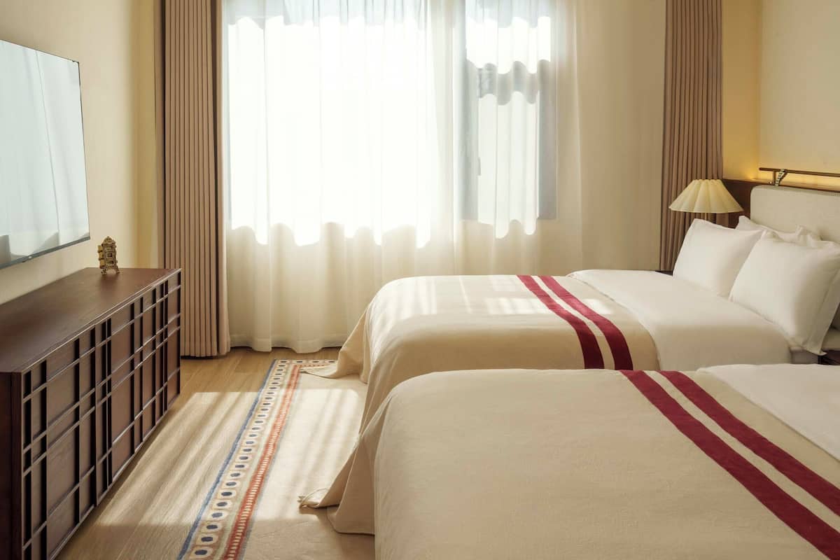 Quarto de hotel com duas camas, uma televisão sobre um móvel de madeira, uma luminária e uma janela com cortinas.