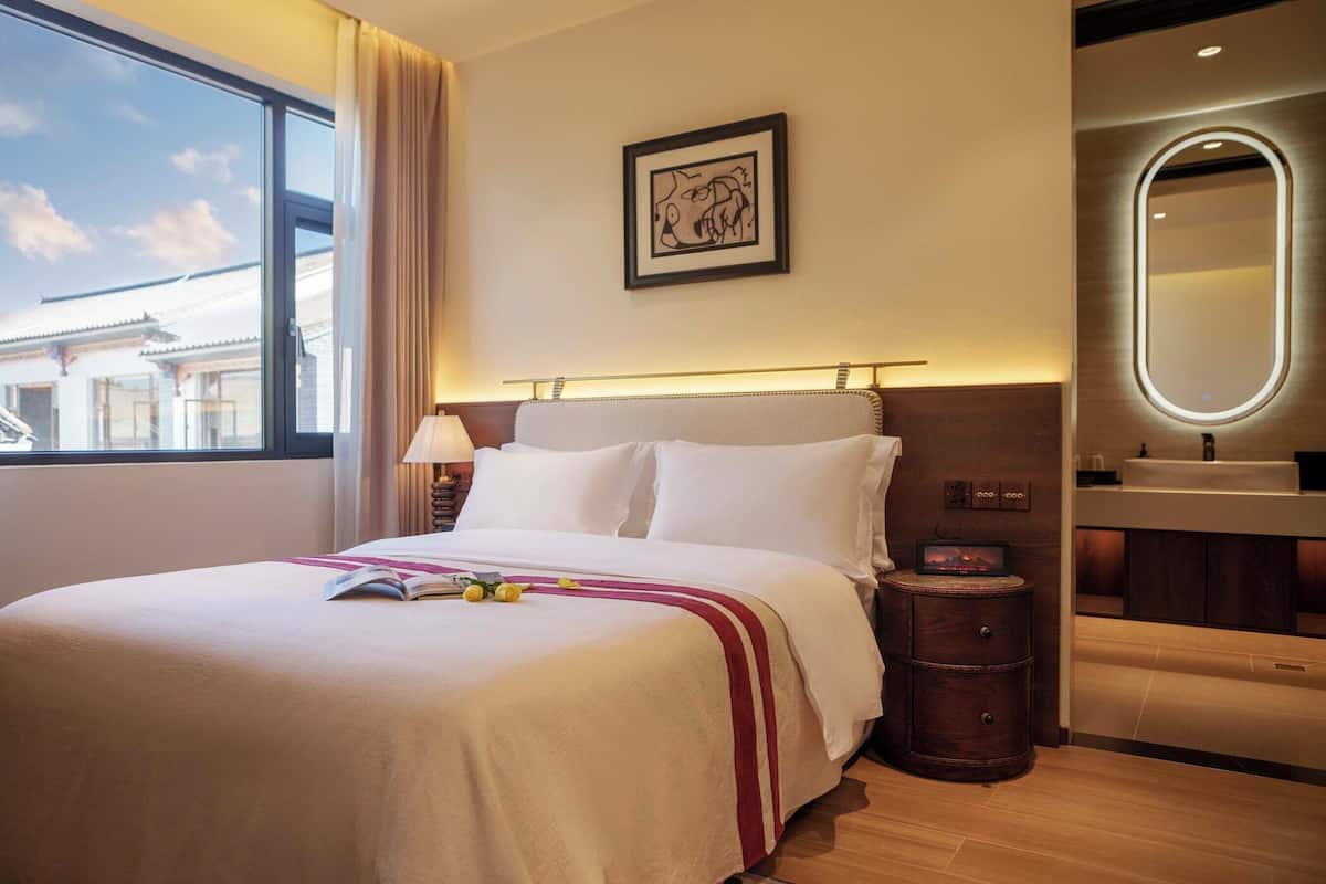 Quarto de hotel com uma cama grande, mesa de cabeceira, abajur e vista para prédios pela janela.