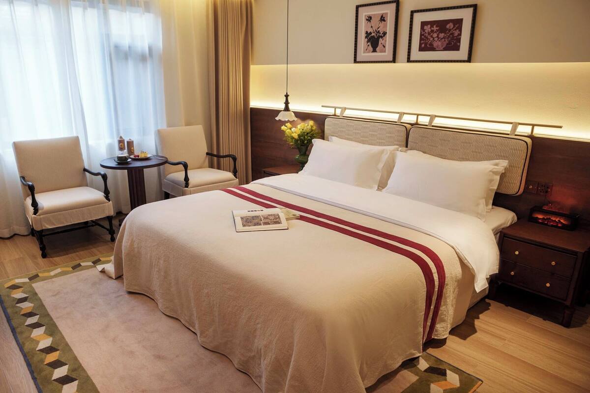 Quarto de hotel com uma cama grande, mesa de cabeceira, uma cadeira, uma mesinha e uma luminária.