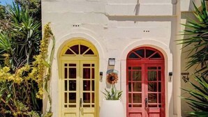 A building with two prominent doors, one yellow and one red, with a large white planter in front. - Gozo Couples Bungalow No 3 · Pool Use & Own Cabana (Gozo)