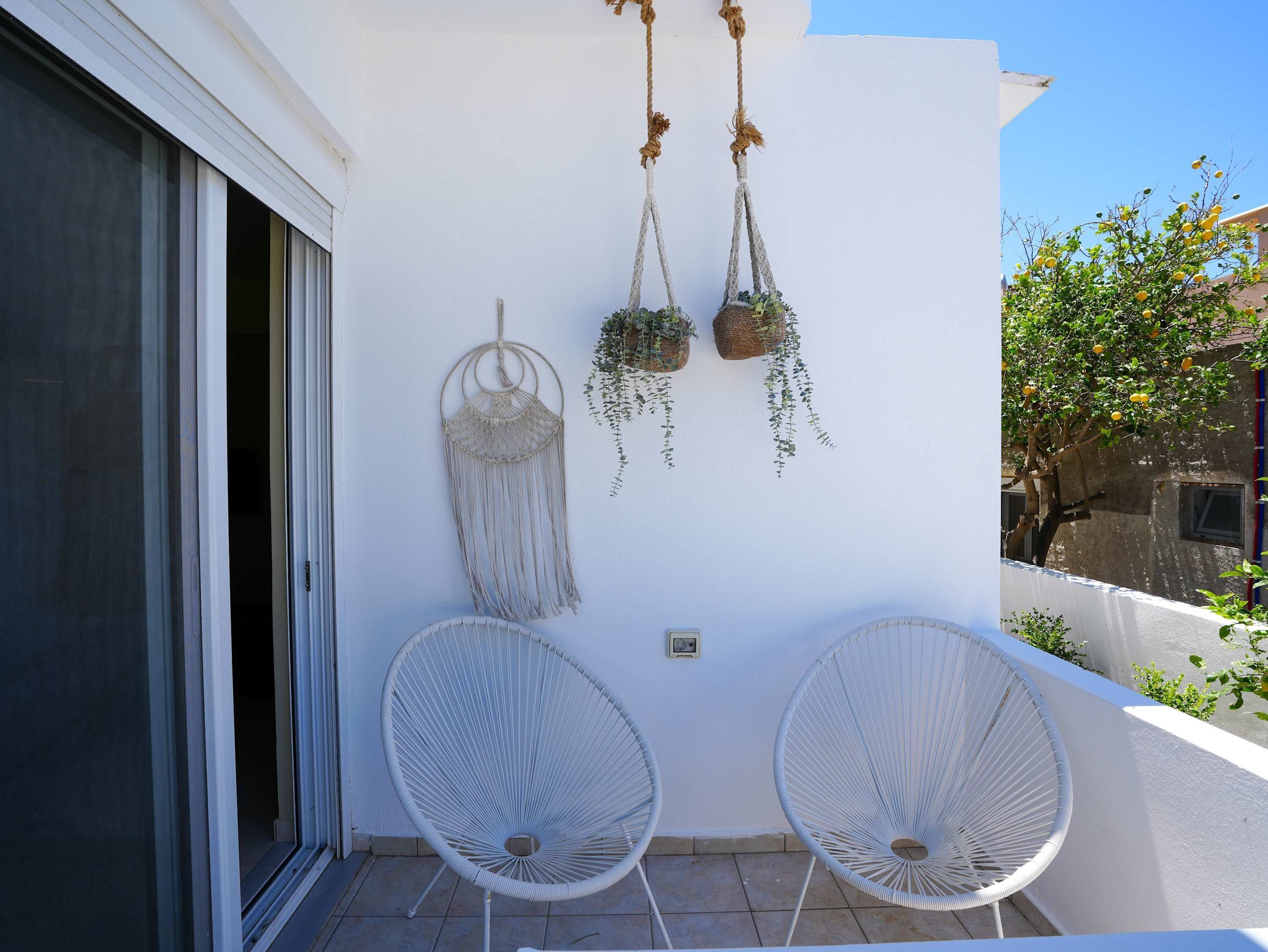 Uma varanda com duas cadeiras brancas de arame, um adorno de macramé na parede e plantas penduradas.