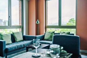 Ein modernes Zimmer mit einem blauen Sofa, einem Tisch mit einem Glas und Blick auf Gebäude und Grünflächen durch große Fenster.