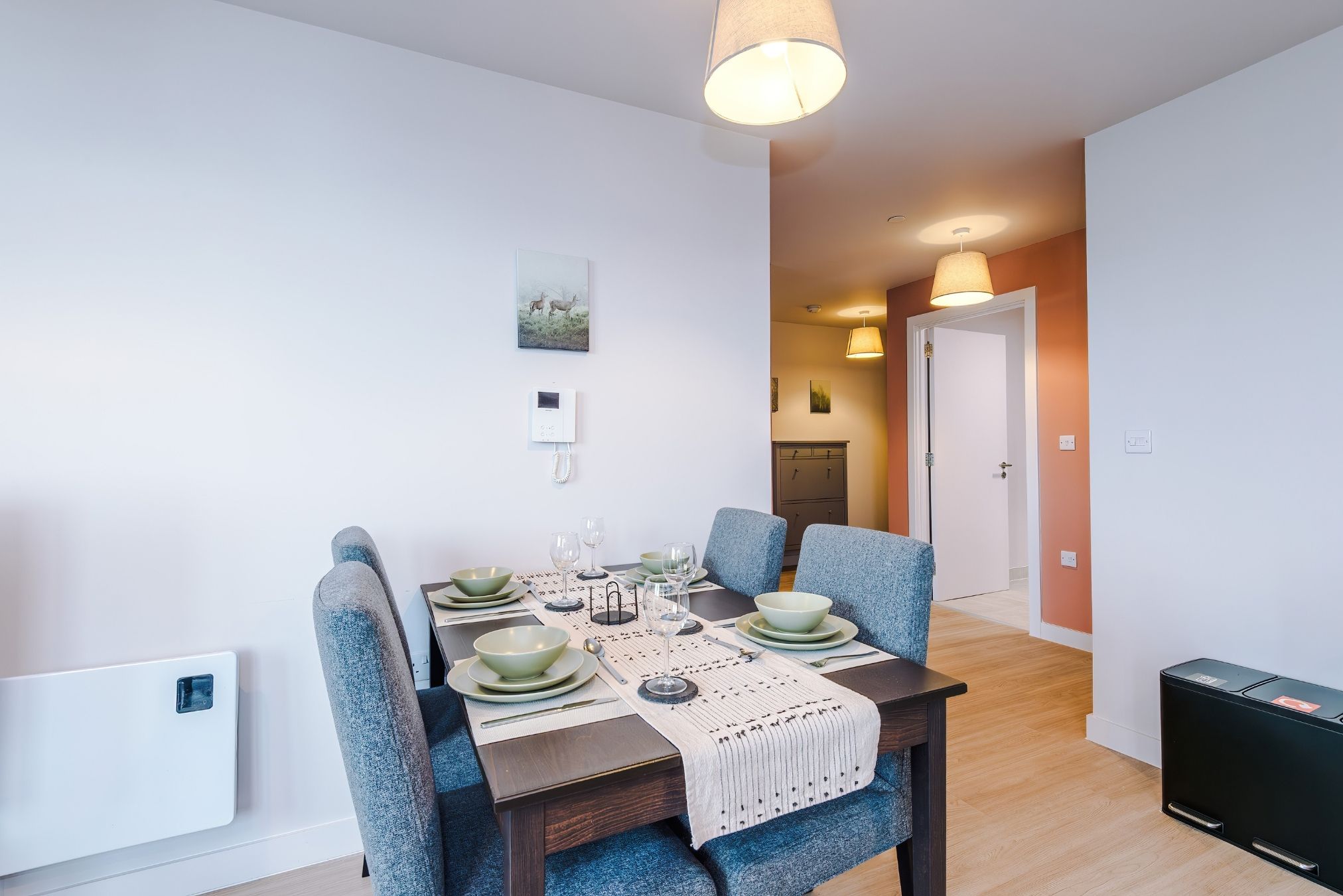 Un comedor con una mesa dispuesta para cuatro, sillas, un teléfono en la pared y una puerta que da a otra habitación.