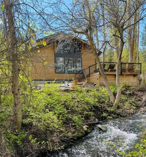 A cabin with a deck overlooking a stream, surrounded by trees and greenery.