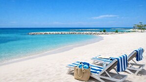 Ein Strand mit weißem Sand, klarem, blauem Wasser und einem Steg. Liegestühle mit blau-weißen gestreiften Handtüchern und einer Strohbag.