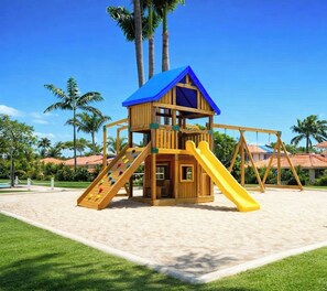 Eine Holzspielanlage mit blauem Dach, Rutschen und einer Kletterwand, umgeben von Sand und Palmen.