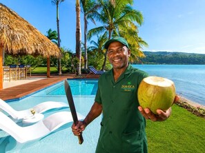 Eine Person hält eine Kokosnuss und ein Messer neben einem Pool mit einem Reetdach und Palmen.