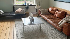 Un salon avec un canapé en cuir marron, une table basse blanche, une grande fenêtre donnant sur un paysage verdoyant et une œuvre d’art encadrée accrochée au mur.