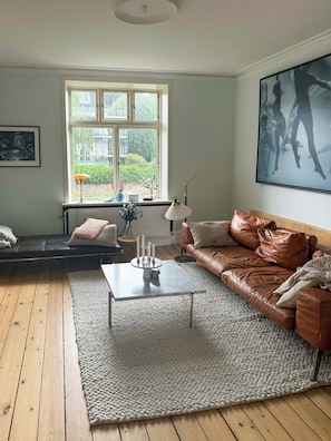 Ein Wohnzimmer mit einem braunen Ledersofa, einem weißen Couchtisch, einem großen Fenster mit Blick auf Grünflächen und einem gerahmten Kunstwerk an der Wand.