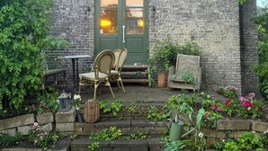 Un bâtiment en briques avec une porte verte, deux fenêtres, un petit porche avec des fauteuils en osier et un jardin où poussent diverses plantes et fleurs.