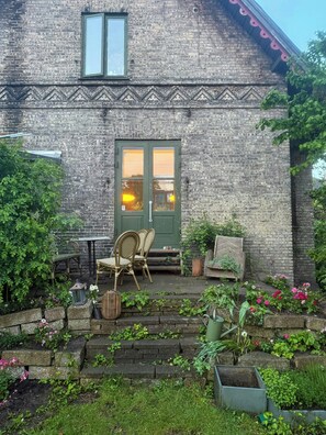 Een bakstenen gebouw met een groene deur, twee ramen, een klein terras met rieten stoelen en een tuin vol planten en bloemen.