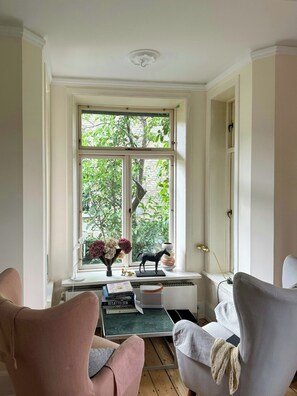 Een kamer met een groot raam, een bureau met boeken en een bloevas, en twee stoelen.