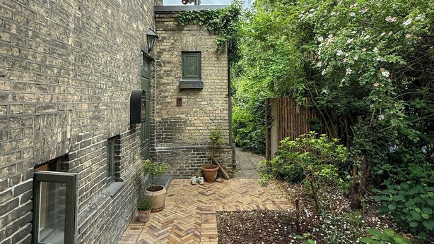 Un bâtiment en briques avec une terrasse dont le sol est pavé selon un motif en chevrons, une petite fenêtre et un pot de fleurs.