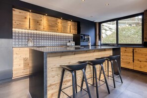 Eine moderne Küche mit Holzschränken, einer sechseckigen Fliesenrückwand, einer integrierten Kochinsel mit Sitzgelegenheiten und einem großen Fenster mit Blick ins Freie.