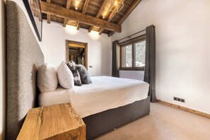 Ein Schlafzimmer mit Bett, Holz-Kopfteil, kleinem Holztisch, Fenster mit Vorhängen und Decke mit sichtbaren Balken.