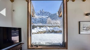 A room with a large window overlooking a snowy mountain landscape, a wooden ceiling, and a television on a wooden cabinet. - Clarines · Where the Fields Meet Mont Blanc (Chamonix)