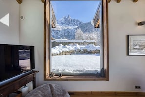 Ein Zimmer mit großem Fenster und Blick auf eine verschneite Berglandschaft, einer Holzbalkendecke und einem Fernseher auf einer Holzkommode.