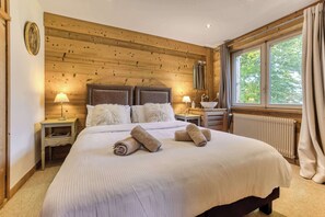 A bedroom with a wooden ceiling, a large bed with white bedding, two bedside tables with lamps, and a window with curtains.