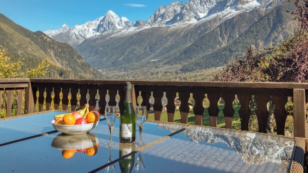 Een tafel met een fruitschaal, een fles en glazen, met uitzicht op een bergachtig landschap.