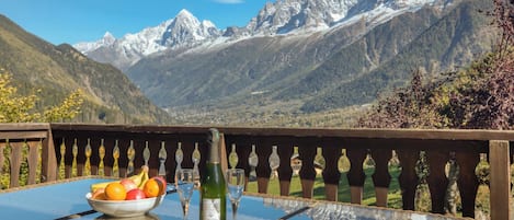 Una mesa con un bol de fruta, una botella y vasos, con vista a un paisaje montañoso.