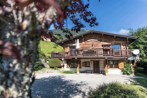 A wooden chalet with a balcony, a gravel driveway, and a mountain backdrop.