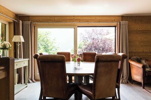 A dining area with a wooden table and leather chairs, a vase with flowers, and a view through large windows.