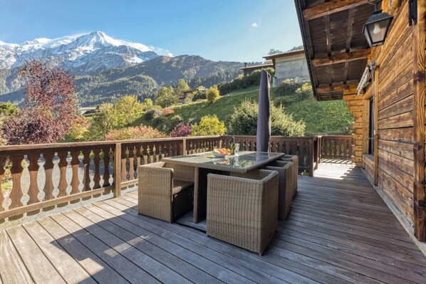 Une terrasse en bois avec une table à plateau de verre et des chaises en osier, offrant une vue sur un paysage montagneux.