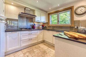 A modern kitchen with white cabinets, a stainless steel range hood, a window with a view of greenery, and a clock on the wall.